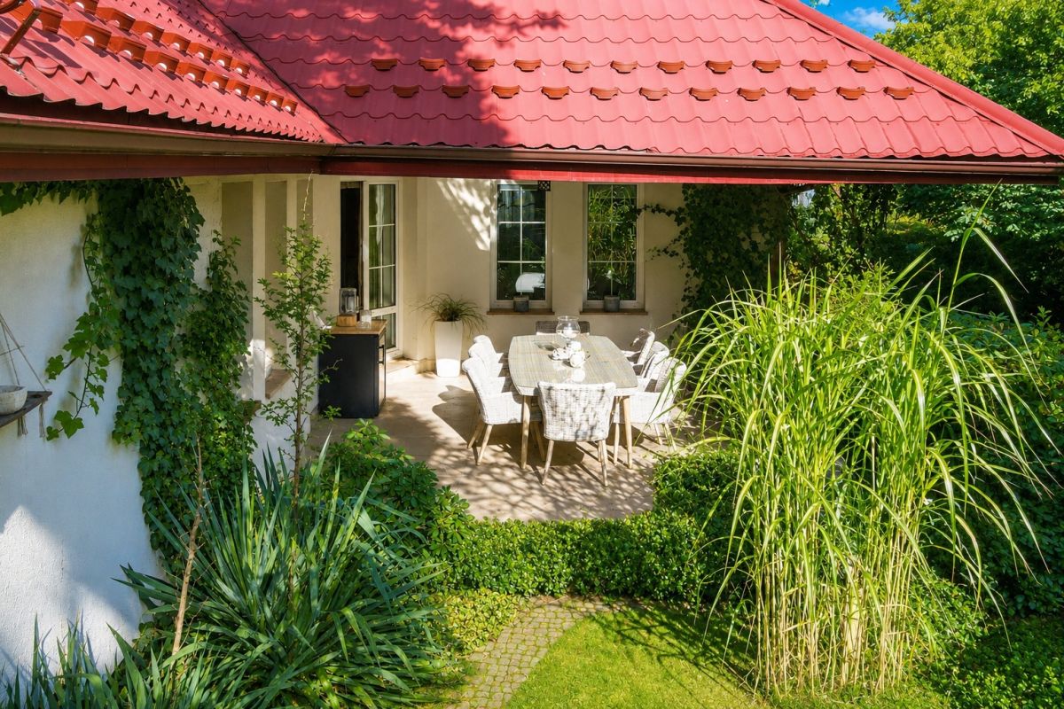 Sunlit patio dining area under a red-tiled roof, with a long table and grey wicker chairs surrounded by lush green plants.