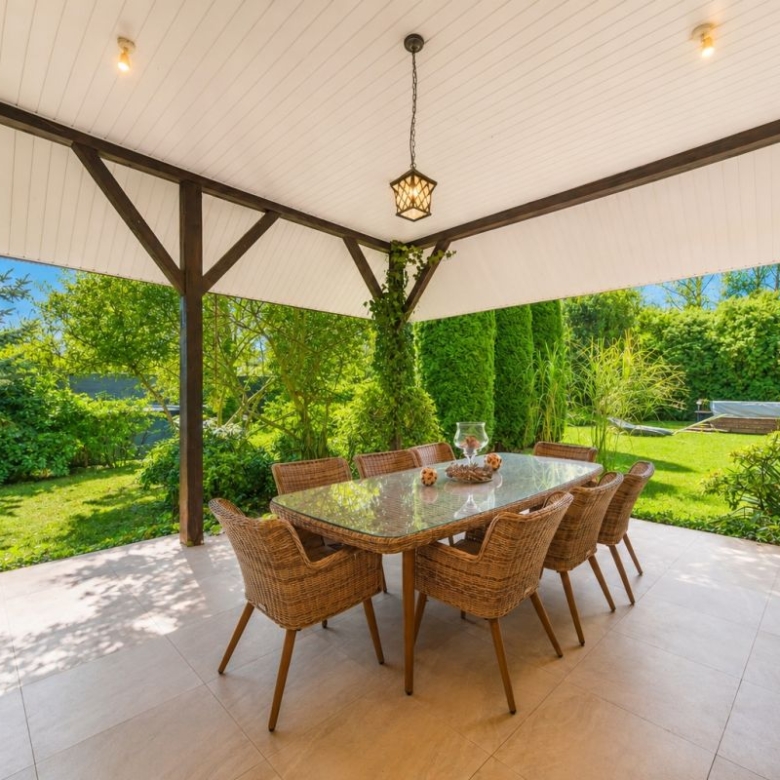 Covered outdoor patio with a glass dining table and wicker chairs overlooking a lush garden under a white ceiling and wooden beams.