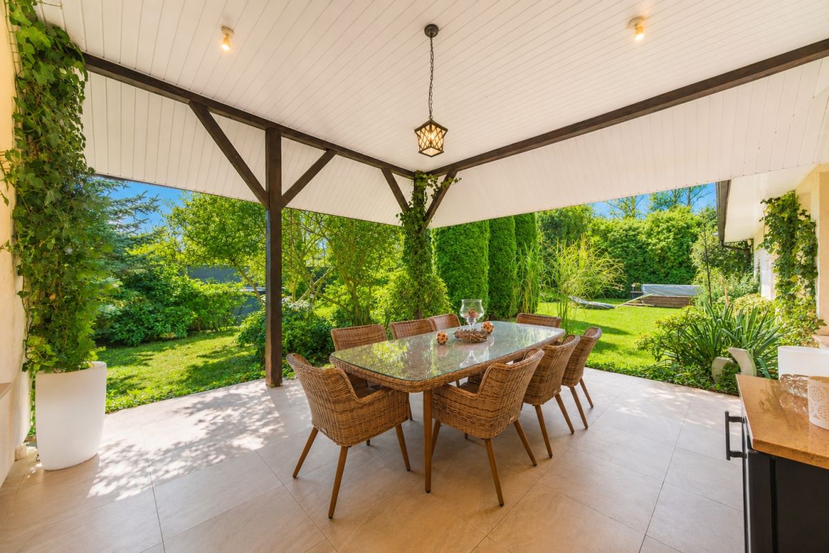 Covered outdoor patio with a glass dining table and wicker chairs overlooking a lush garden under a white ceiling and wooden beams.