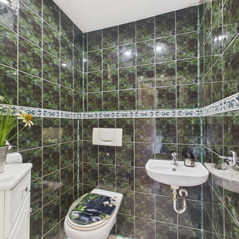 Green marble-tiled bathroom with a white toilet and wall-mounted sink; decorative floral seat cover and a potted plant on a white cabinet sits in the corner.