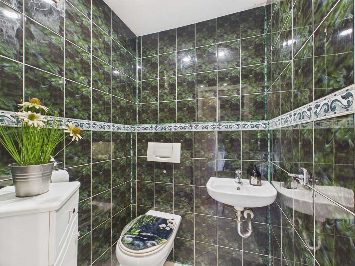 Green marble-tiled bathroom with a white toilet and wall-mounted sink; decorative floral seat cover and a potted plant on a white cabinet sits in the corner.