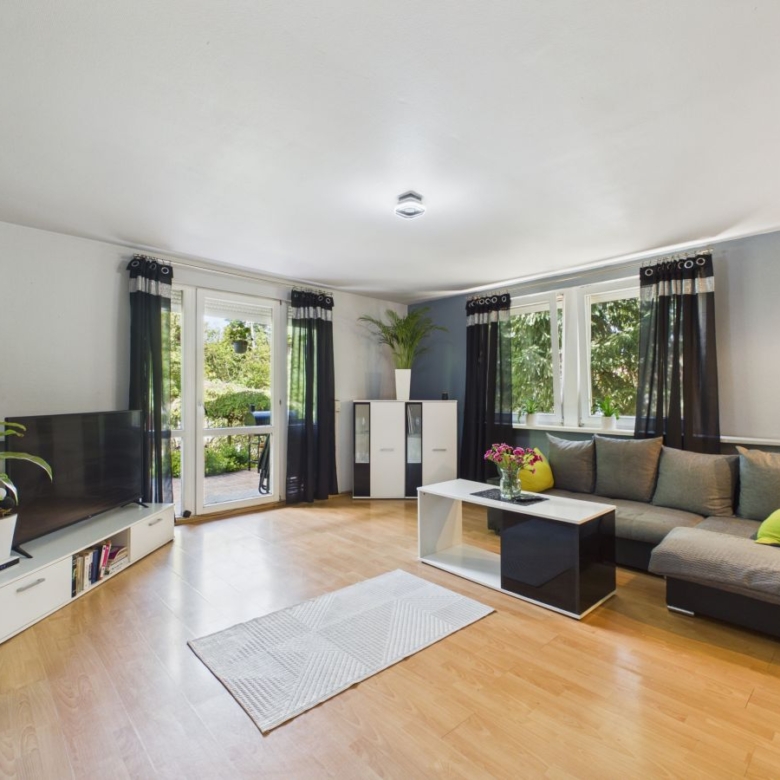 Bright living room with a gray sectional sofa, white coffee table, and a TV on a low white stand near large windows with black curtains.