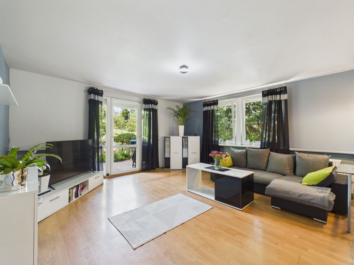 Bright living room with a gray sectional sofa, white coffee table, and a TV on a low white stand near large windows with black curtains.