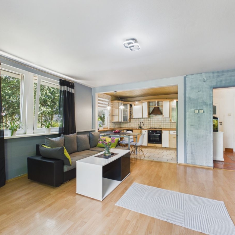 Open-plan living area with a gray sofa, white coffee table, and large windows; kitchen seen in background.