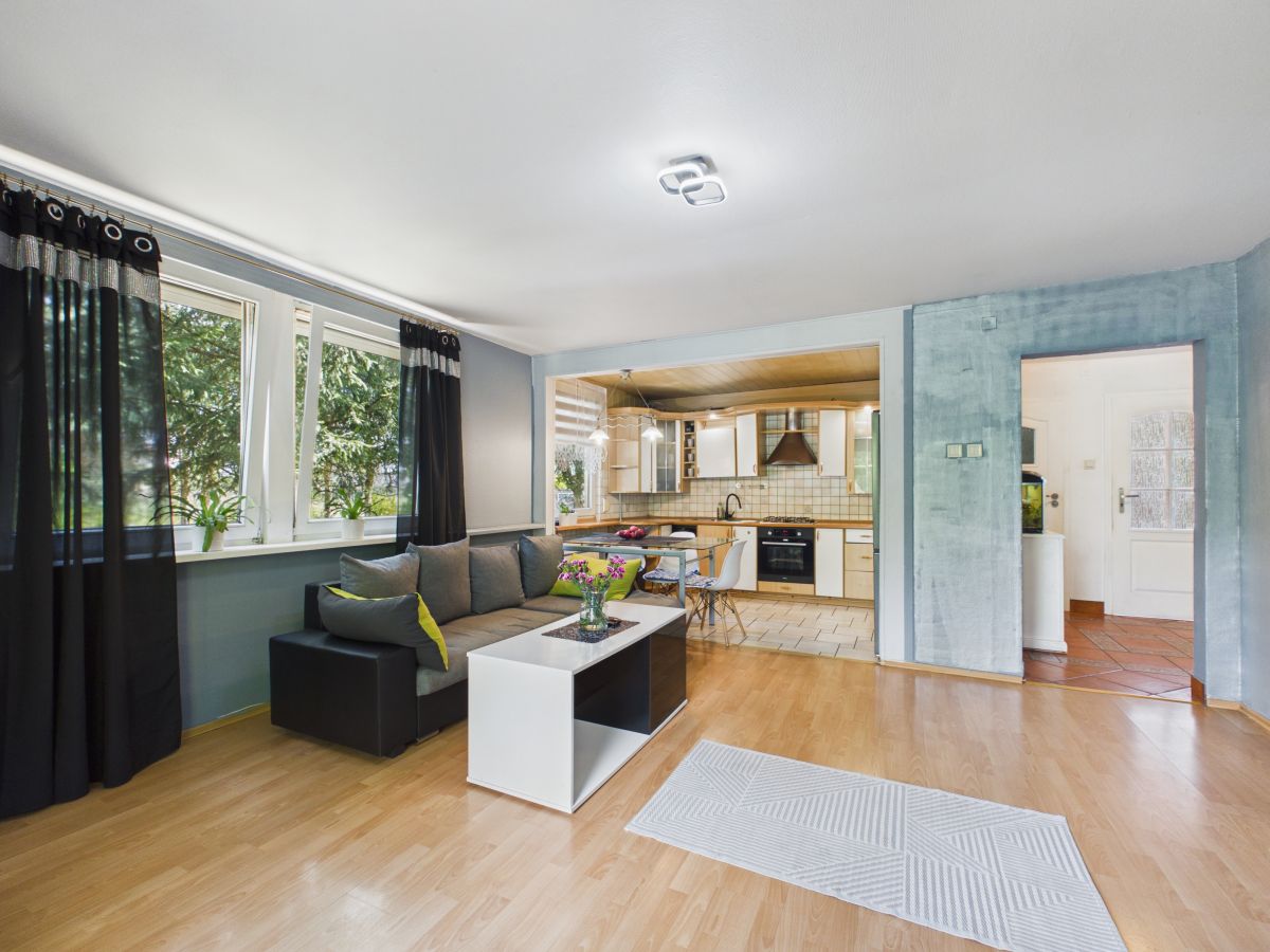 Open-plan living area with a gray sofa, white coffee table, and large windows; kitchen seen in background.