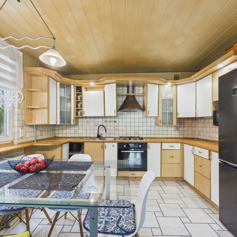 Bright kitchen with light wood cabinets, tiled backsplash, stainless steel fridge, and glass dining table.