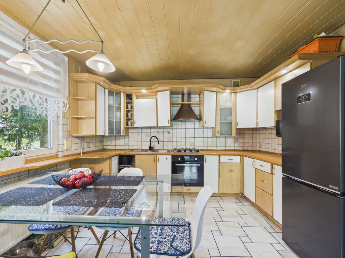 Bright kitchen with light wood cabinets, tiled backsplash, stainless steel fridge, and glass dining table.