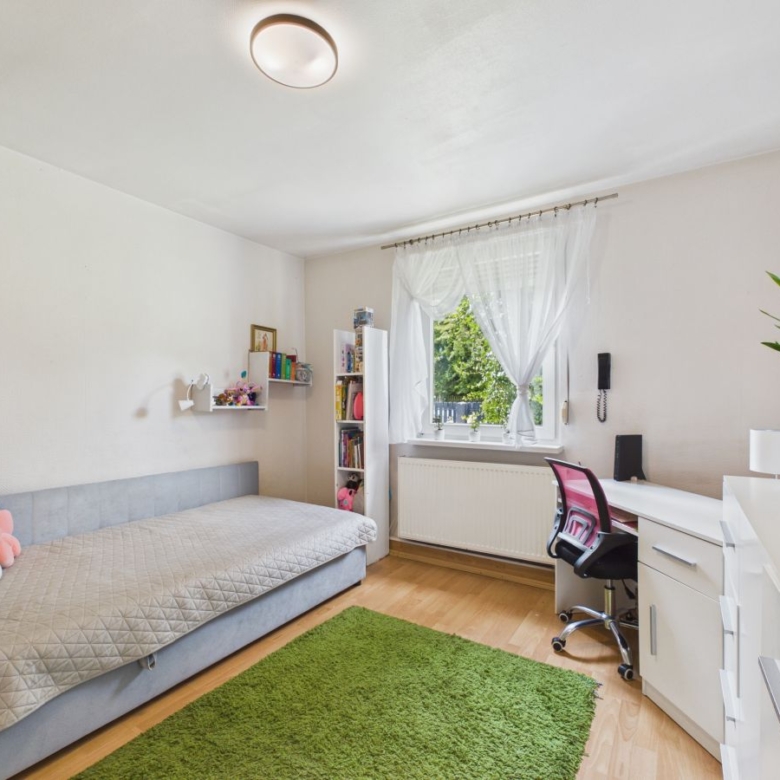 Bright, tidy bedroom with a light gray daybed along the left wall, a green shag rug, and a white desk setup by a window with sheer curtains. A pink and black office chair sits at the desk, a tall white bookshelf and floating shelves hold books and decor, and a potted plant sits on the dresser to the right.
