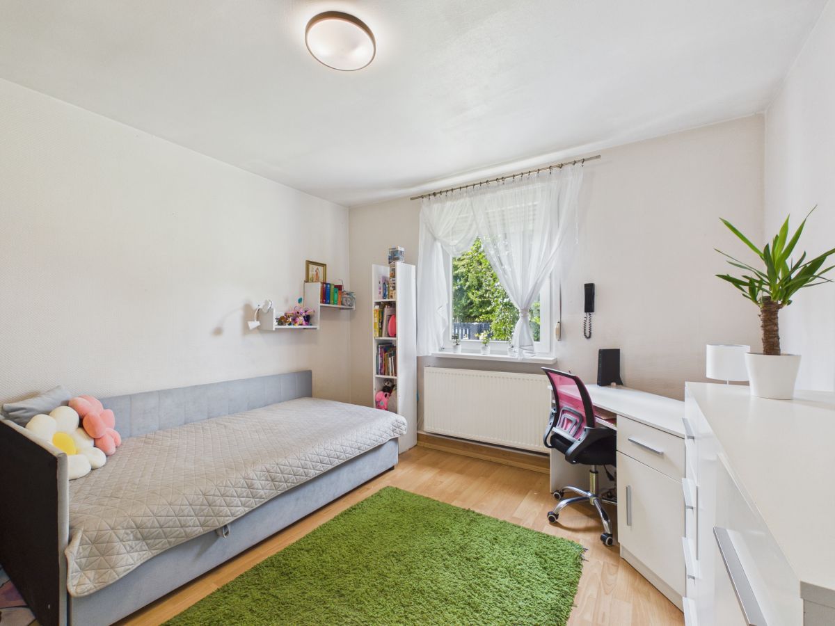 Bright, tidy bedroom with a light gray daybed along the left wall, a green shag rug, and a white desk setup by a window with sheer curtains. A pink and black office chair sits at the desk, a tall white bookshelf and floating shelves hold books and decor, and a potted plant sits on the dresser to the right.