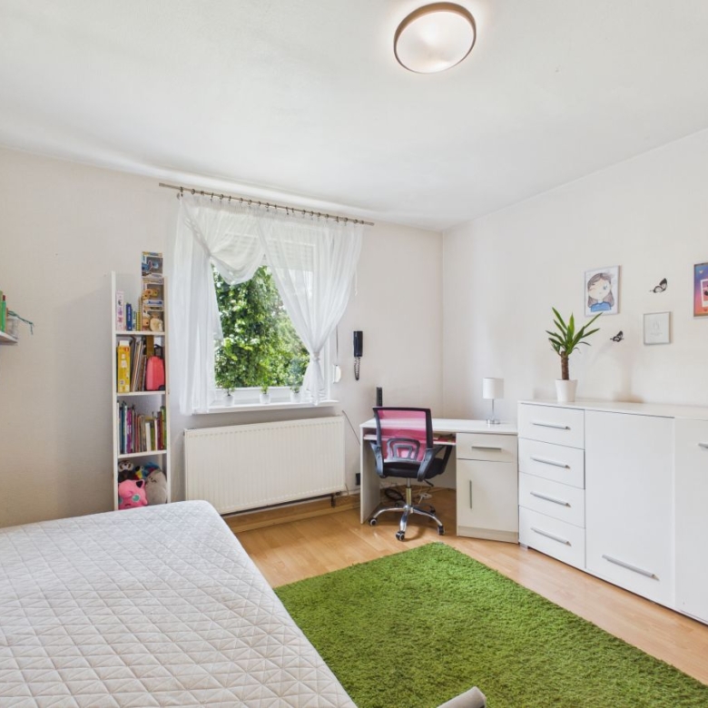 Bright, tidy bedroom with a white quilted bed, green rug, and white furniture including a desk and dresser by the window. A pink chair sits at the desk under sheer curtains.