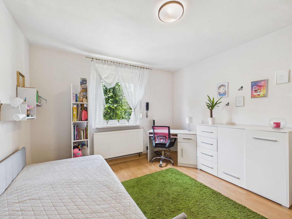 Bright, tidy bedroom with a white quilted bed, green rug, and white furniture including a desk and dresser by the window. A pink chair sits at the desk under sheer curtains.