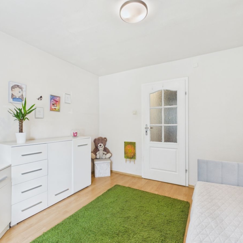 Bright kid’s bedroom with white dresser and desk, a potted plant, teddy bear, and a green rug on a light wood floor.