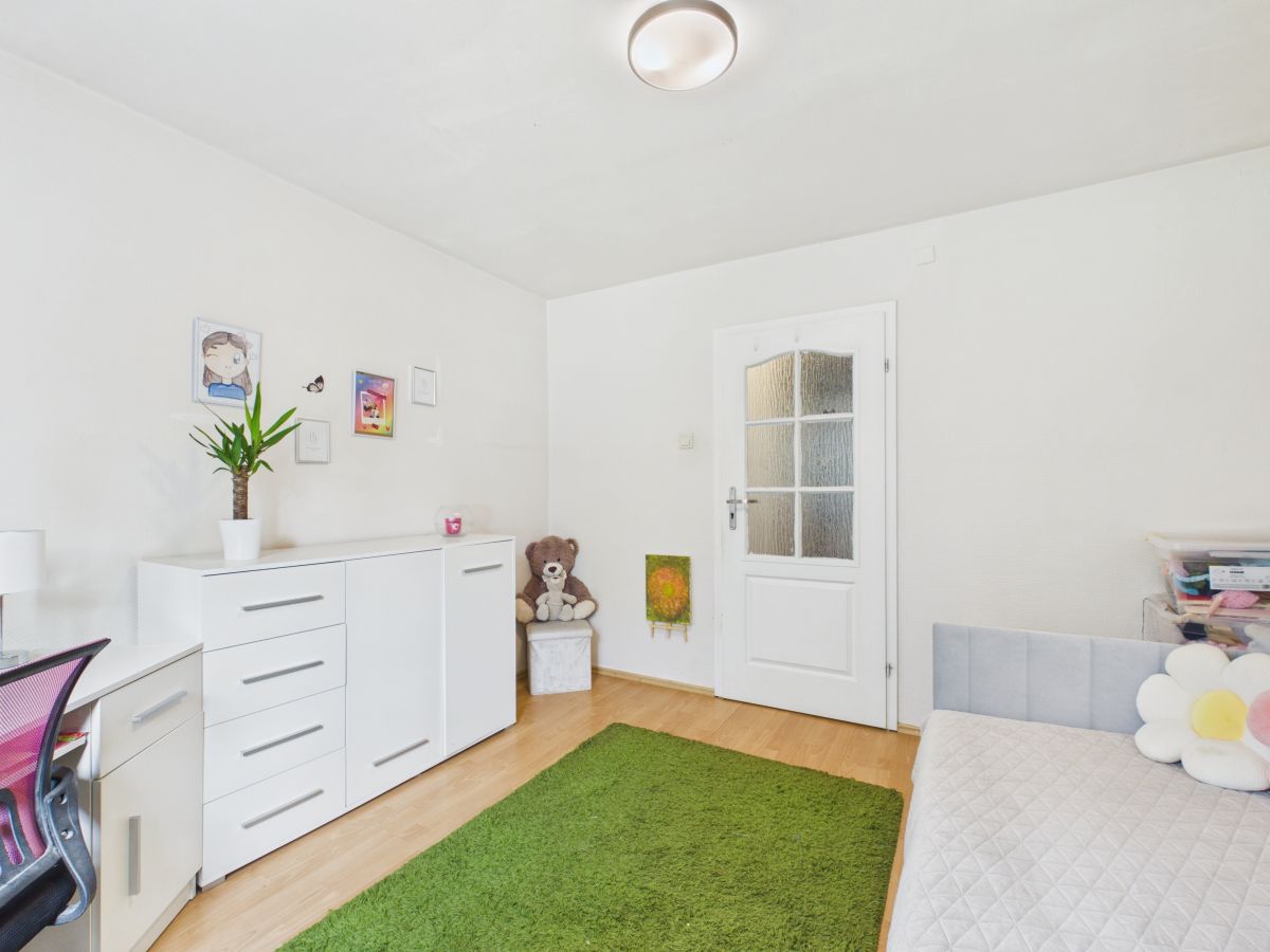 Bright kid’s bedroom with white dresser and desk, a potted plant, teddy bear, and a green rug on a light wood floor.