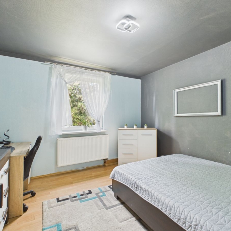 A small modern bedroom with a bed, white dresser, and a desk along light blue walls and a window with sheer curtains over a radiator on the left.