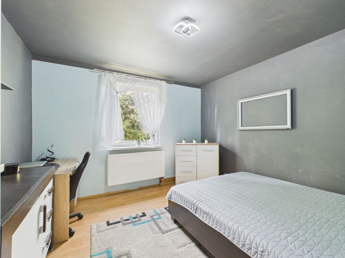A small modern bedroom with a bed, white dresser, and a desk along light blue walls and a window with sheer curtains over a radiator on the left.