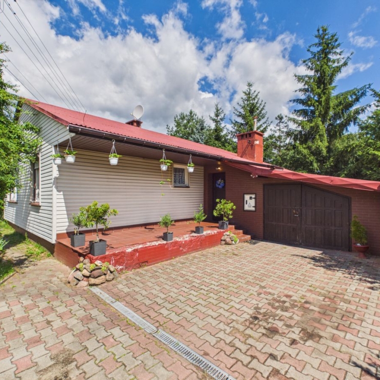 Front view of a single-story house with a red roof and brick garage, surrounded by potted plants on a paved driveway.
