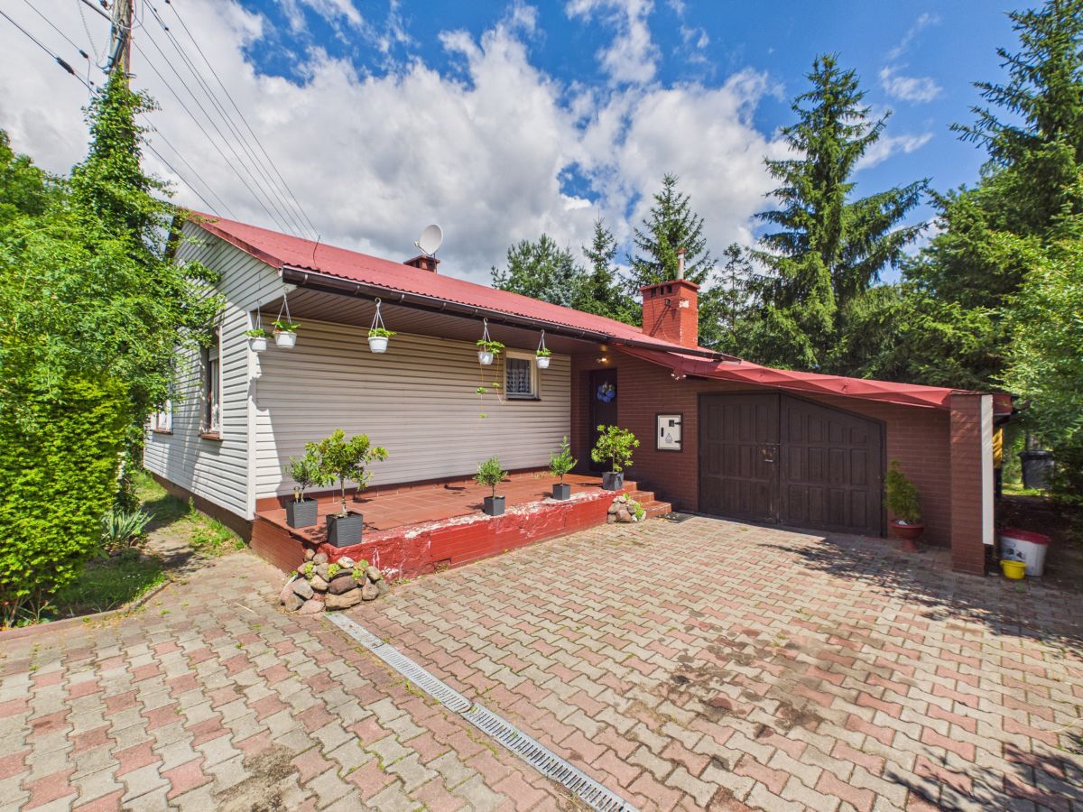 Front view of a single-story house with a red roof and brick garage, surrounded by potted plants on a paved driveway.