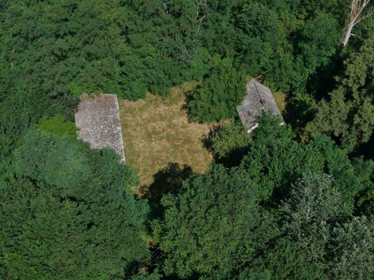 Aerial view of a small grassy clearing with two old stone buildings partially hidden by dense green trees.