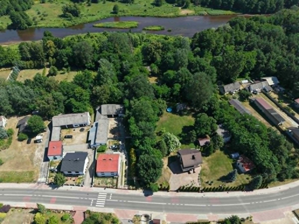 Aerial view of a small neighborhood along a curved road, with red- and gray-roofed houses and dense trees, and a river in the background.