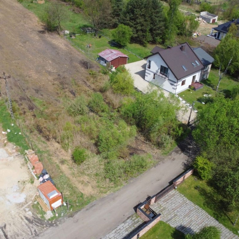 Aerial view of a suburban scene with a white two-story house, green trees, and a dirt road running beside the property.