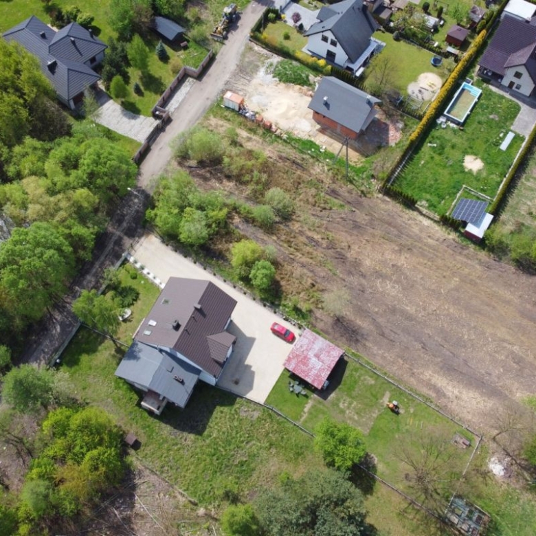 Aerial view of a suburban neighborhood with green trees, houses, and a large empty brown field next to a fenced yard and a red car in the driveway.