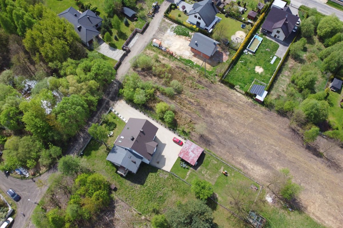 Aerial view of a suburban neighborhood with green trees, houses, and a large empty brown field next to a fenced yard and a red car in the driveway.