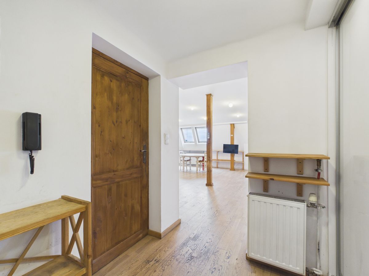 Bright hallway with a wooden door on the left, wall-mounted phone, bench, and shelving; open living area ahead.
