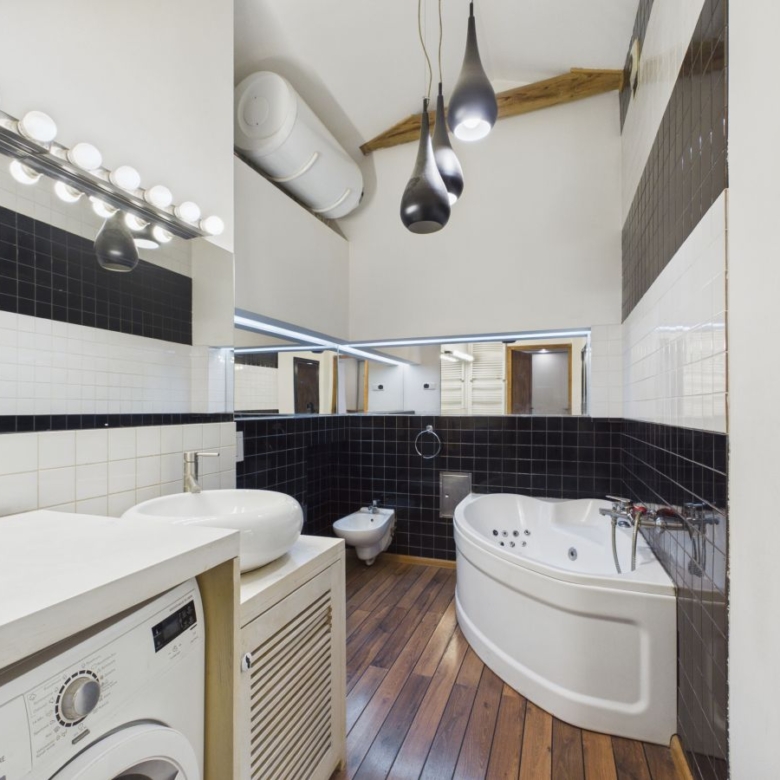 Modern bathroom with a curved white corner bathtub, washing machine, and white vessel sink on a wooden floor with black and white tiled walls.