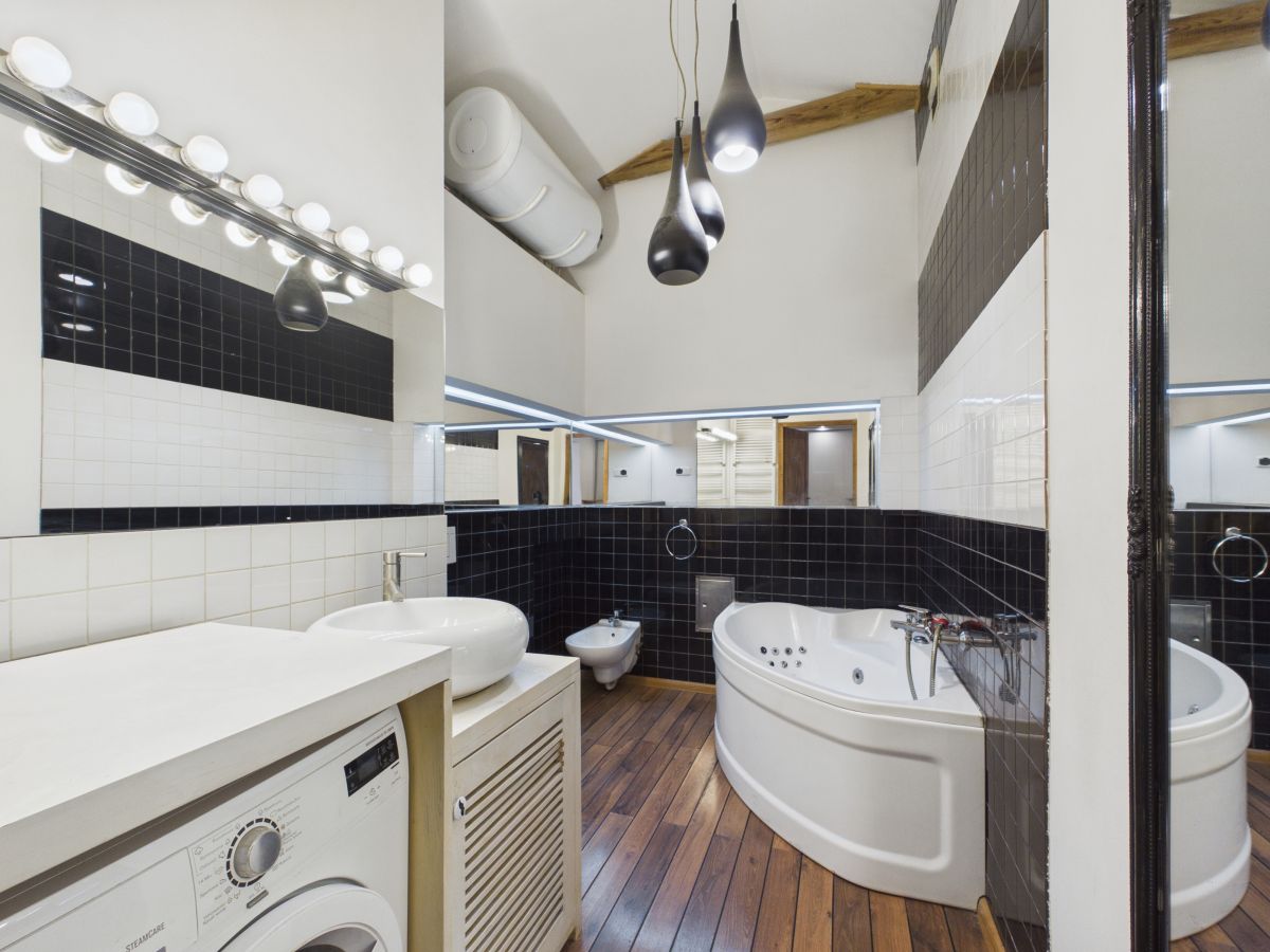 Modern bathroom with a curved white corner bathtub, washing machine, and white vessel sink on a wooden floor with black and white tiled walls.
