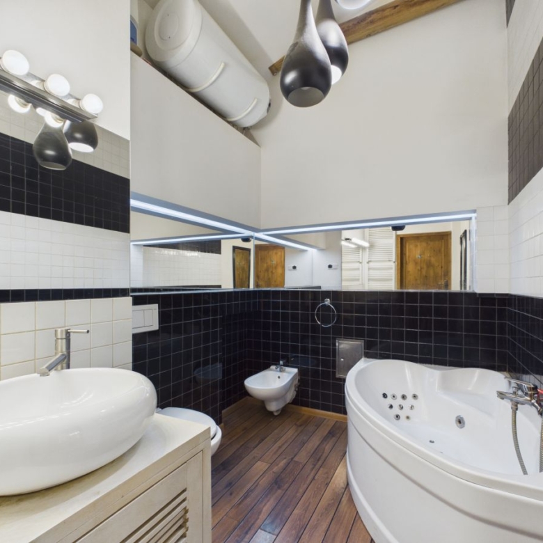 Modern bathroom with a large corner whirlpool tub, white vessel sink, bidet, and wood plank floor; black-and-white tiled walls and a long mirror reflect overhead lighting.