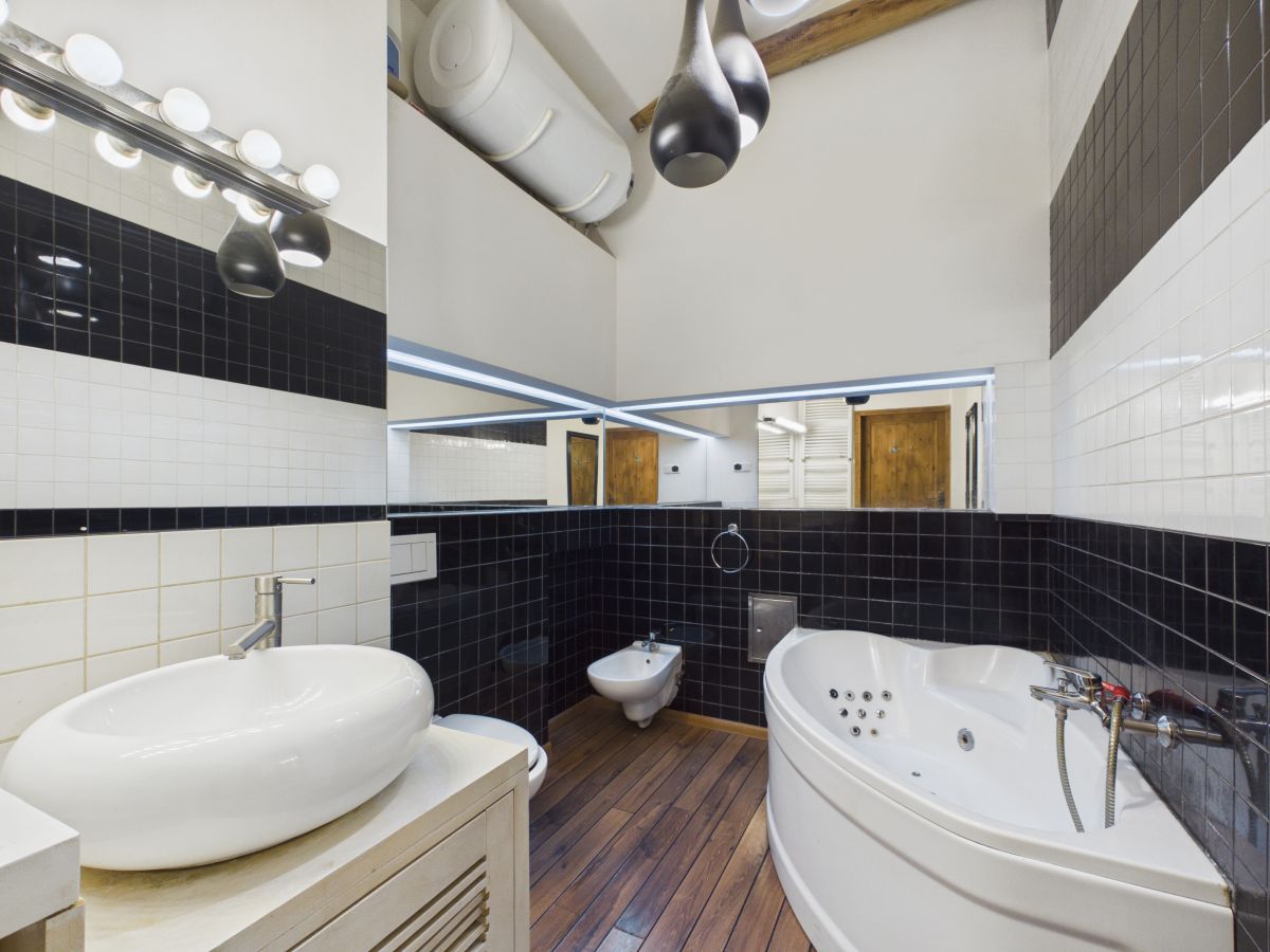 Modern bathroom with a large corner whirlpool tub, white vessel sink, bidet, and wood plank floor; black-and-white tiled walls and a long mirror reflect overhead lighting.