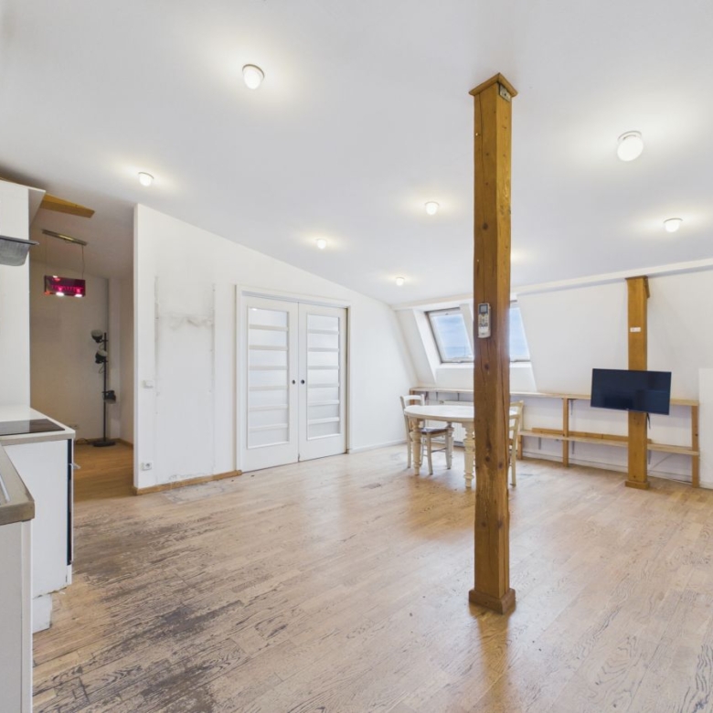Spacious loft with wooden beams, kitchen on left, dining table near a skylight, and a TV on the right wall in a bright, white-walled room.