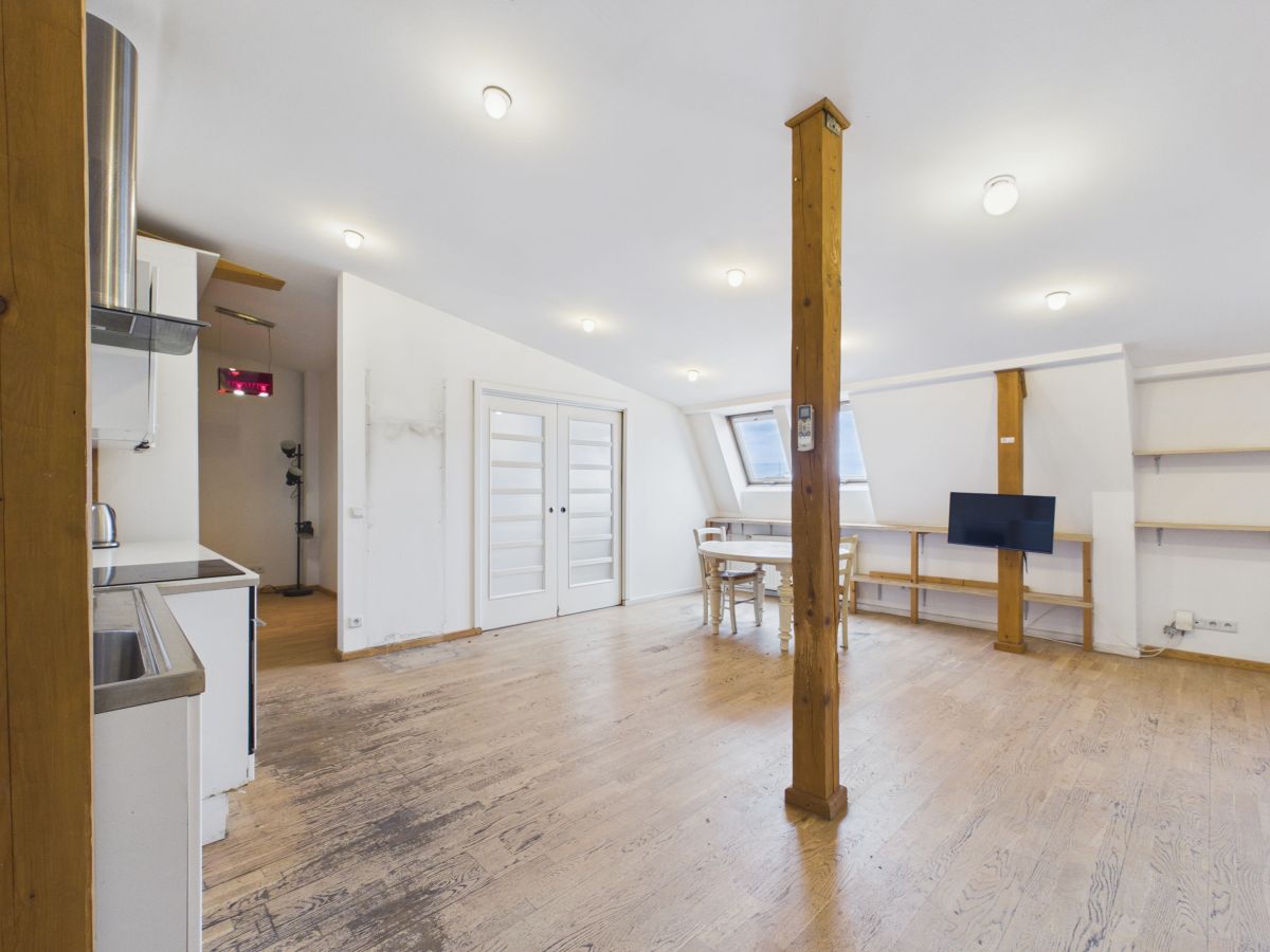 Spacious loft with wooden beams, kitchen on left, dining table near a skylight, and a TV on the right wall in a bright, white-walled room.