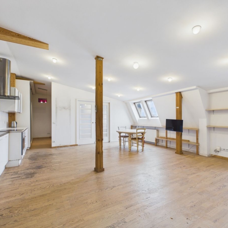 Bright open loft with kitchen on the left, wooden floors, and exposed beams; a round dining table near skylight windows ensures natural light.