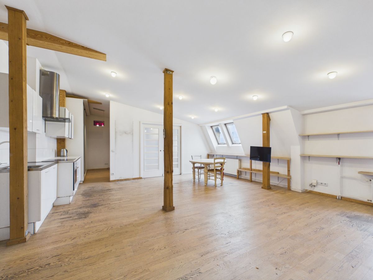 Bright open loft with kitchen on the left, wooden floors, and exposed beams; a round dining table near skylight windows ensures natural light.