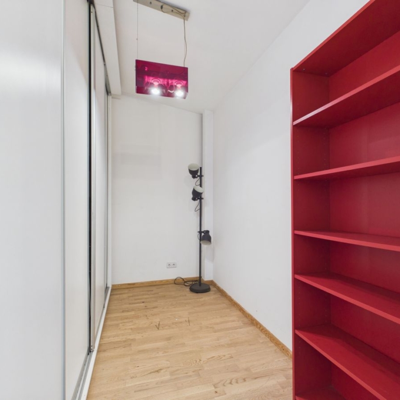 Narrow white-walled room with light wood floor, a red open bookshelf on the right, and a tall black floor lamp near the back; glass sliding door on the left.