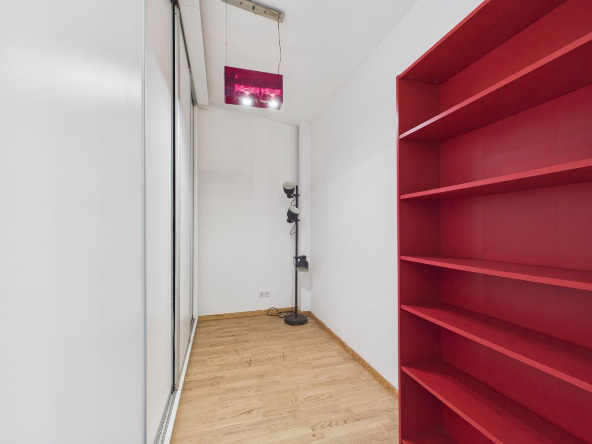 Narrow white-walled room with light wood floor, a red open bookshelf on the right, and a tall black floor lamp near the back; glass sliding door on the left.