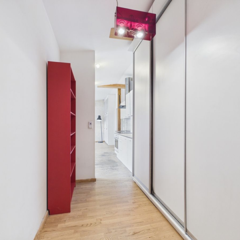 Narrow hallway with light wood floor, red shelving unit on the left and white sliding closet doors on the right, leading to a kitchen."”