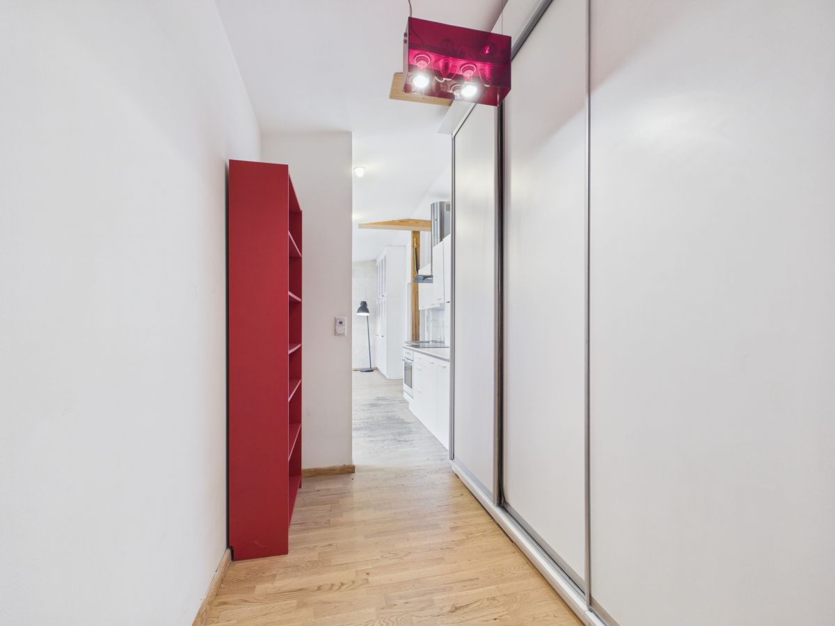 Narrow hallway with light wood floor, red shelving unit on the left and white sliding closet doors on the right, leading to a kitchen."”