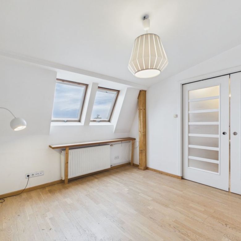 Bright attic room with two skylight windows, white walls, wooden floor, radiator, and a long desk under the skylights near a wooden beam.
