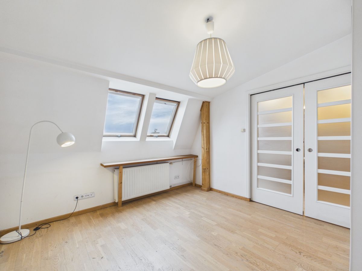 Bright attic room with two skylight windows, white walls, wooden floor, radiator, and a long desk under the skylights near a wooden beam.