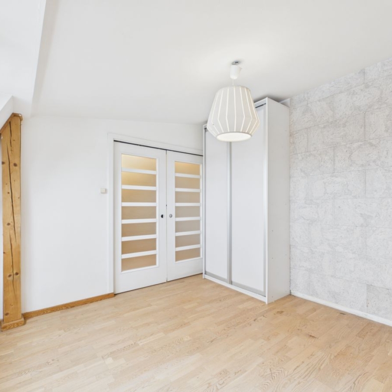 Bright minimalist attic room with white walls, a wooden beam, desk, and glass-panel double doors beside a tall white wardrobe.