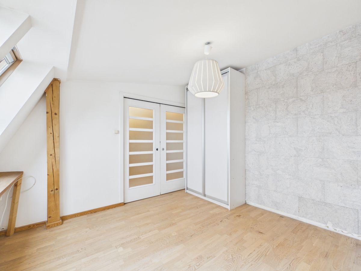 Bright minimalist attic room with white walls, a wooden beam, desk, and glass-panel double doors beside a tall white wardrobe.