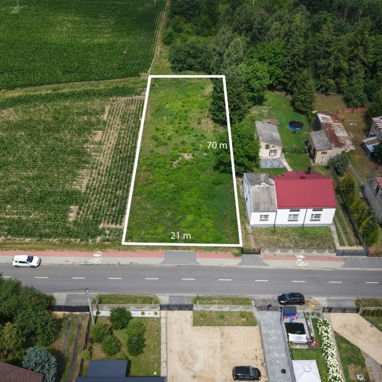 Aerial view of a rectangular vacant lot outlined in white, measuring 70 m by 21 m, adjacent to a residential street and green fields nearby.