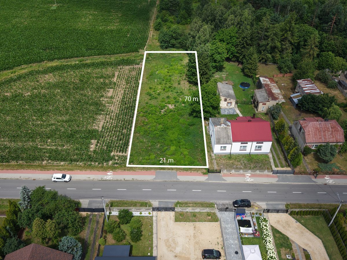 Aerial view of a rectangular vacant lot outlined in white, measuring 70 m by 21 m, adjacent to a residential street and green fields nearby.