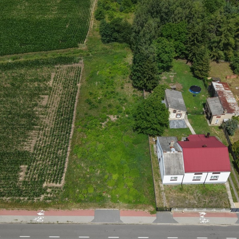 Aerial view of a rural property with a white house and red roof, several outbuildings, and a fenced yard next to green fields and trees.