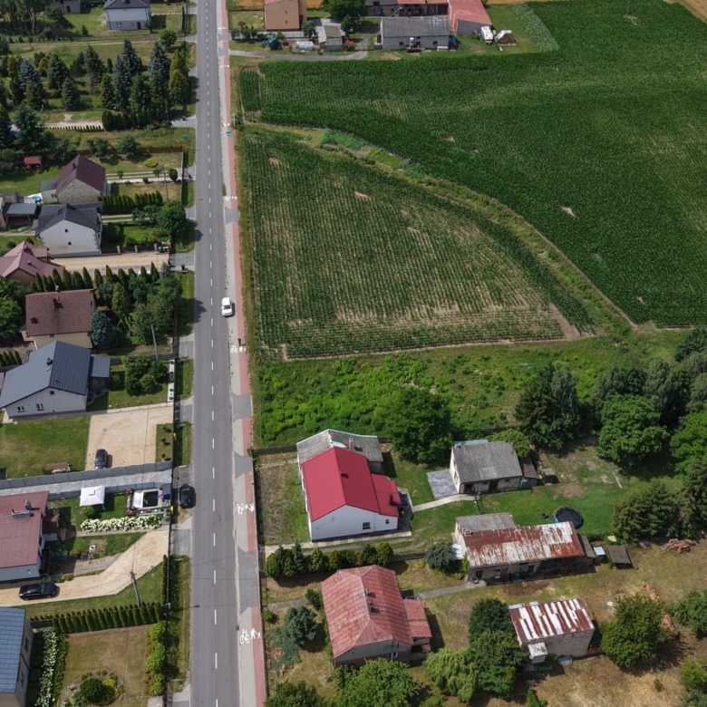 Aerial view of a rural street with houses on the left and green fields on the right, showing a central road and surrounding homes and farmland.