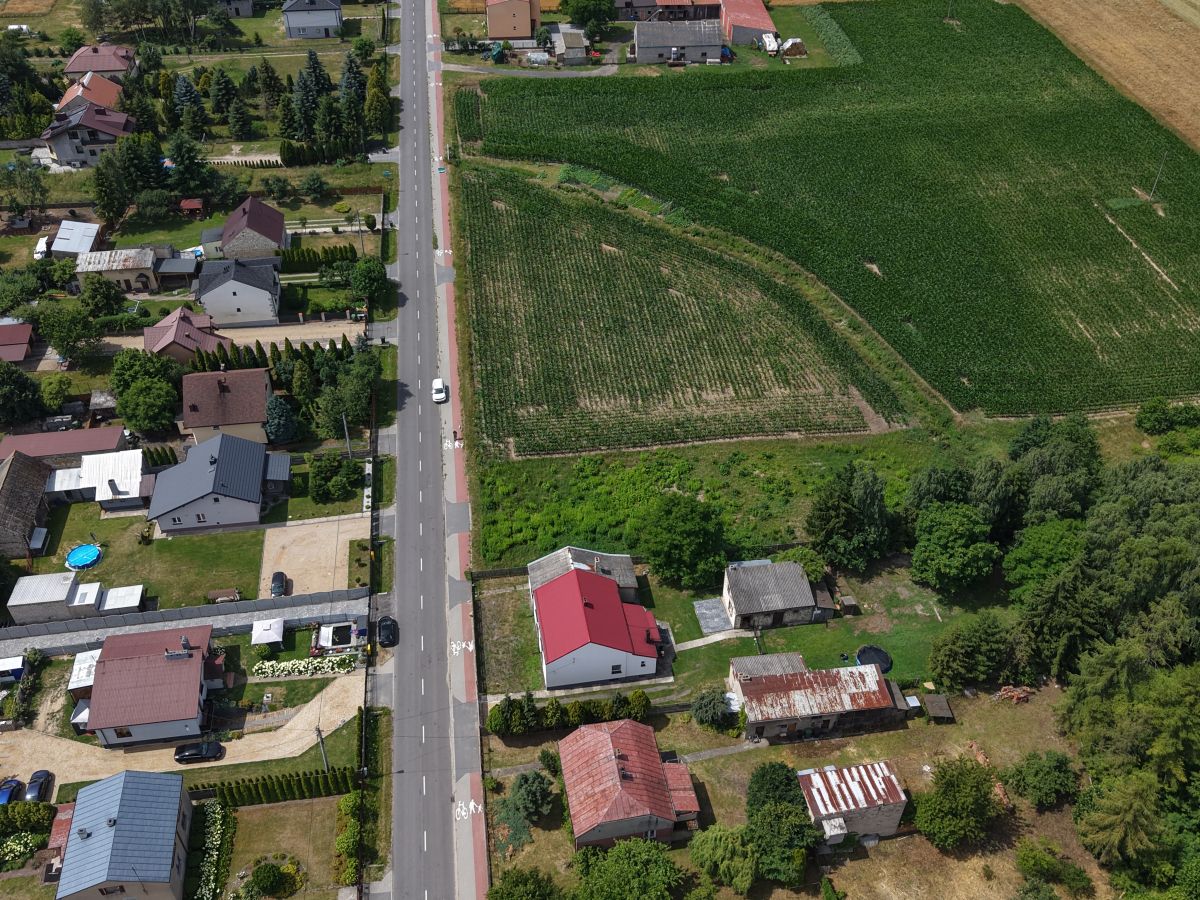 Aerial view of a rural street with houses on the left and green fields on the right, showing a central road and surrounding homes and farmland.