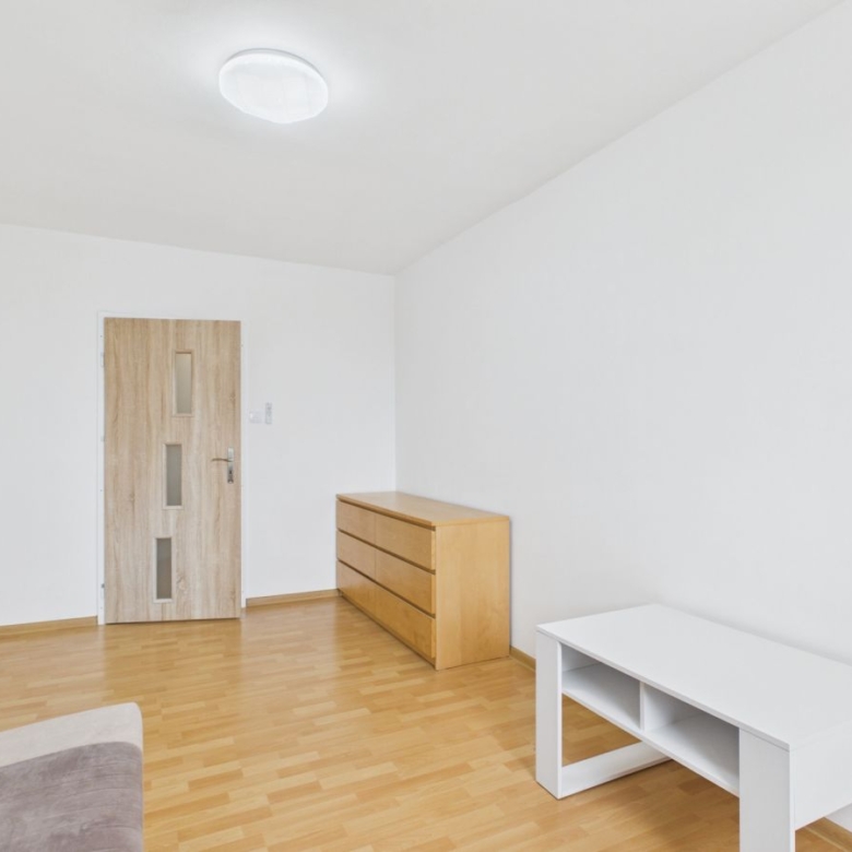 Minimalist living room with light wood floor, white walls, a wooden door, a three‑drawer dresser, and a white storage table; sofa edge visible in foreground.
