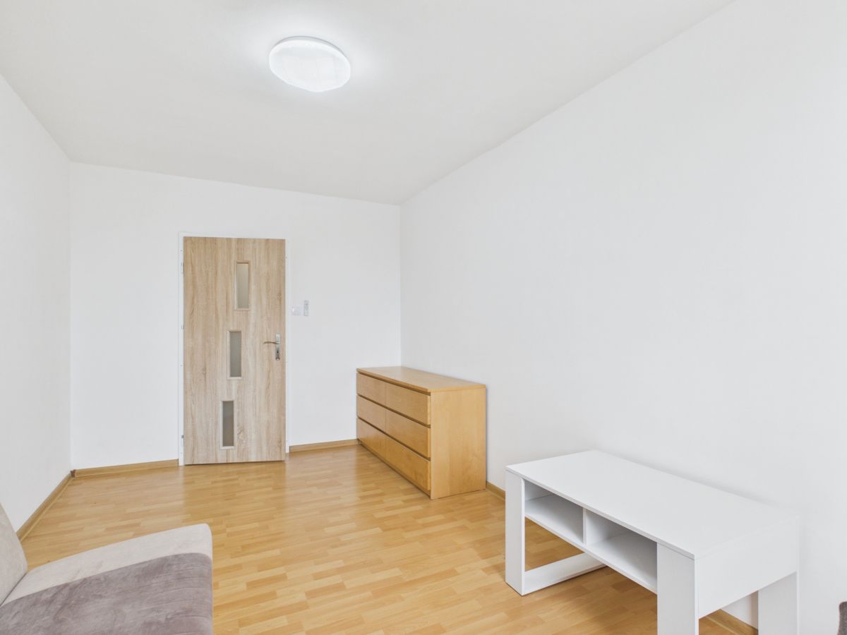 Minimalist living room with light wood floor, white walls, a wooden door, a three‑drawer dresser, and a white storage table; sofa edge visible in foreground.